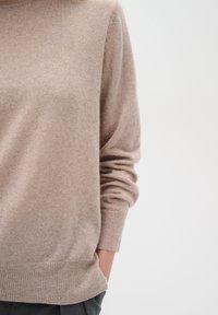 Pull en tricot beige clair à coupe décontractée, avec des poignets et un ourlet côtelés. Texture douce et manches l�égèrement amples, porté sur un pantalon gris anthracite.