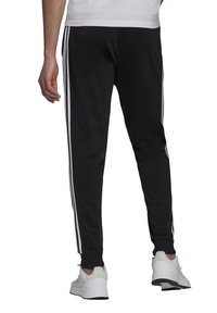 Schwarze Sporthose mit weißen Seitenstreifen, elastischem Bund und gerippten Knöcheln, kombiniert mit weißen Sneakers. Glatte Textur, lässiges Design.