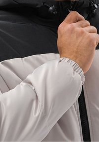 Lightweight, padded jacket with a black hood and gray body. Features elastic cuffs and a zippered front for secure closure.