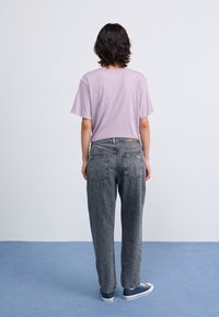 Jeans de mezclilla gris con un corte relajado y cinco bolsillos, combinados con una camiseta morada clara, llevados con zapatillas navy sobre un suelo azul.