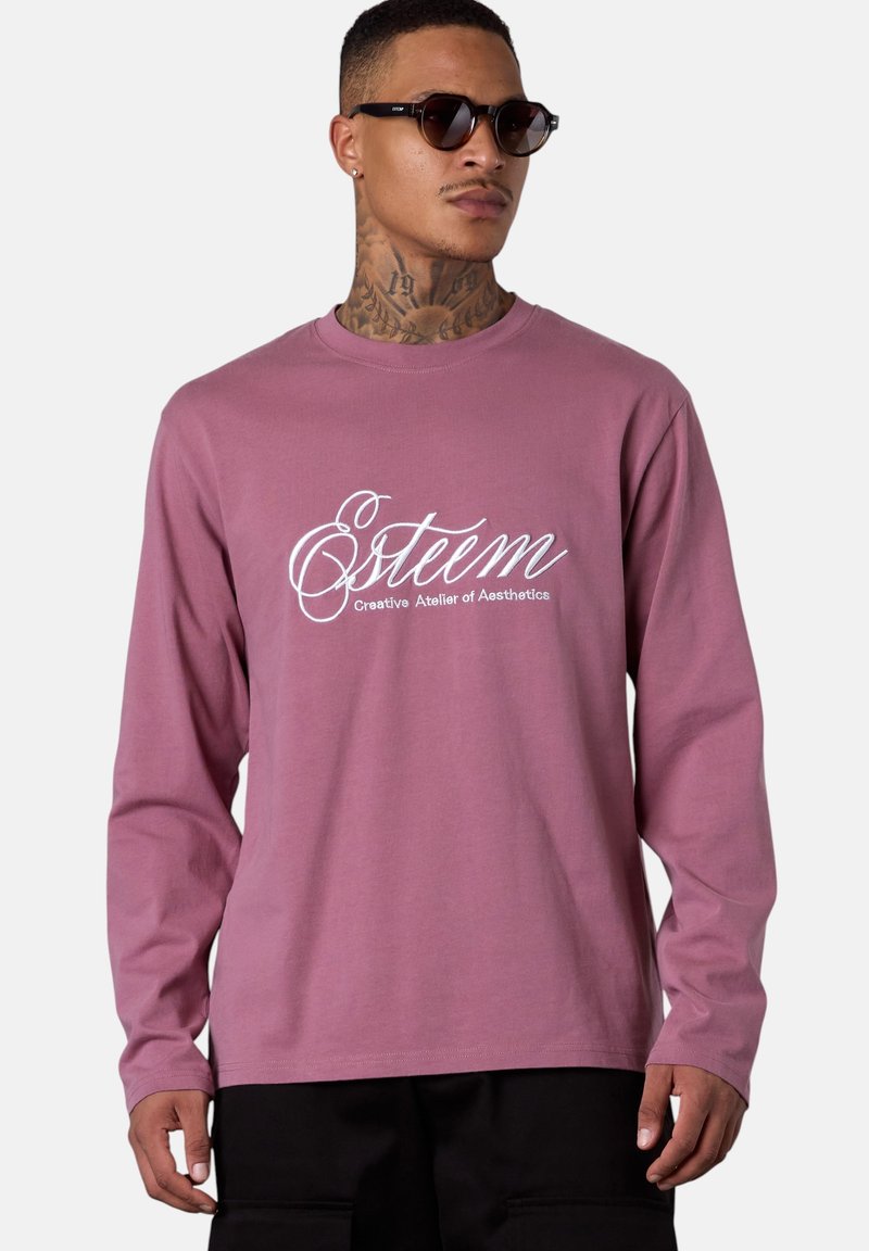 Mann trägt ein lila langärmliges Hemd mit dem Aufdruck "Esteem Creative Atelier of Aesthetics", schwarze Hose, Sonnenbrille und einen Nackentattoo vor einem neutralen Hintergrund.