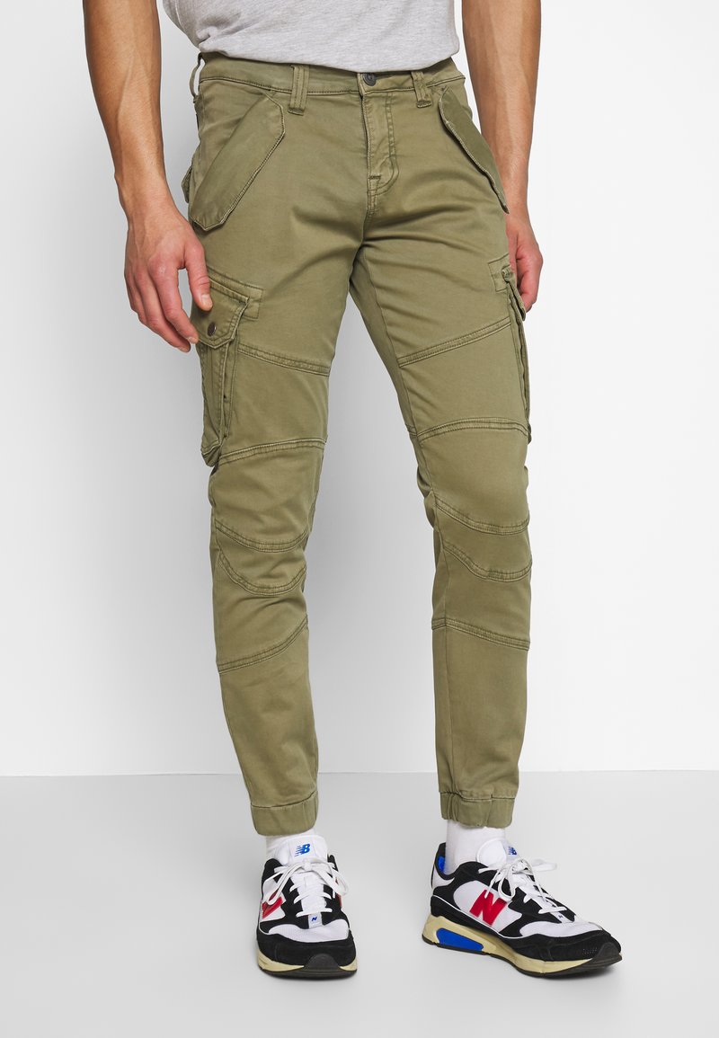 Olijfgroene cargobroek met zijzakken, slim fit ontwerp en gestikte panelen. Gecombineerd met zwart-witte sneakers met rode en blauwe accenten.