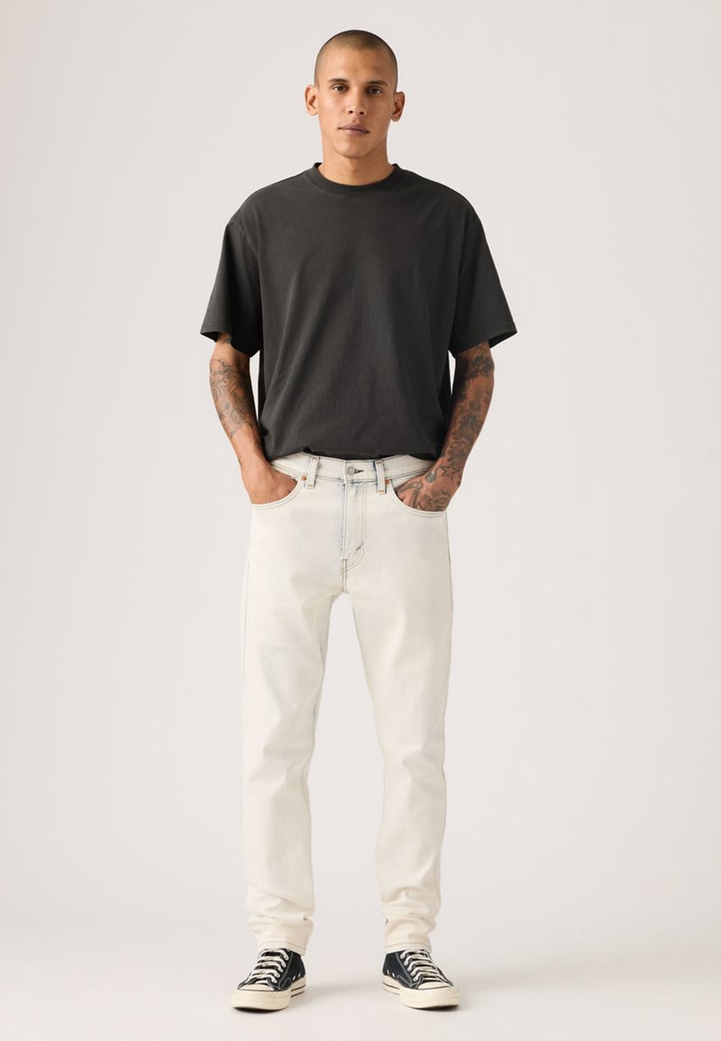 T-shirt noir à col rond, jean en denim clair et baskets noires. Le mannequin a des tatouages et pose les mains dans les poches. Design simple et décontracté.