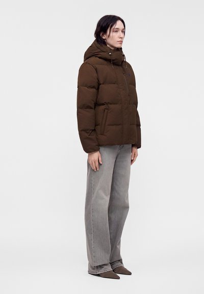 Femme debout portant une doudoune marron avec capuche, un jean gris large et des chaussures pointues devant un fond clair uni.