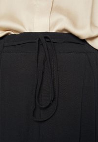 Haut beige rentré dans un pantalon noir avec un fin cordon en tissu noir noué lâchement à la taille.