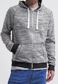 Uomo che indossa una felpa grigia e nera con zip e lacci bianchi, jeans blu e un tatuaggio visibile sul collo e sulla mano.