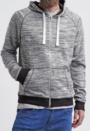 Man wearing a gray and black zip-up hoodie with white drawstrings, blue jeans, and a visible tattoo on his neck and hand.