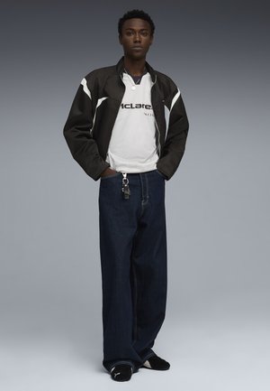 Giovane in piedi con le mani in tasca, indossa una giacca da corsa bianca e nera, una maglietta bianca McLaren, jeans scuri larghi e scarpe nere Puma.