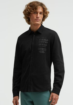 Junger Mann mit lockigem Haar, der ein schwarzes Hemd mit Knöpfen trägt, auf der Brust befindet sich ein heller Schriftzug, und eine türkise Hose, stehend vor einem einfarbigen Hintergrund.