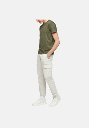 Mann trägt olivgrünes kurzärmeliges T-Shirt, hellbeige Cargohose mit Taschen, weiße Socken und weiße Turnschuhe, steht mit den Händen in den Taschen.