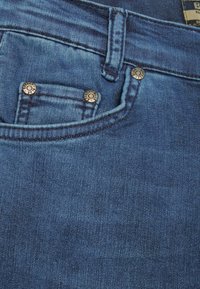 Jeans de mezclilla azul con una cintura de media altura, que presentan un pequeño bolsillo frontal, remaches metálicos y una textura suave.