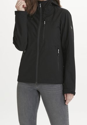 Femme portant une veste noire zippée avec capuche et un jean gris, main près du col, debout devant un fond blanc uni.