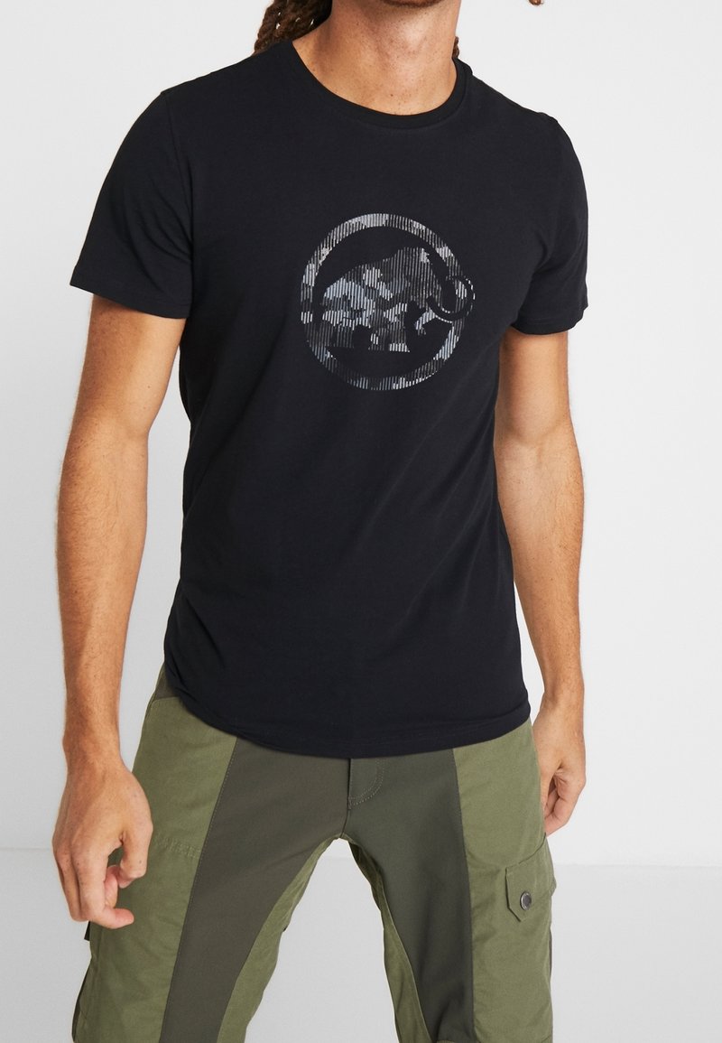 Mann trägt schwarzes T-Shirt mit rundem abstraktem Tierdesign und zweifarbigen grünen Cargohosen, steht mit entspannten Armen.