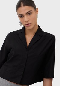 Mujer joven con el cabello peinado hacia atrás, lleva pendientes plateados en forma de disco y una camisa negra con cuello y mangas de tres cuartos.
