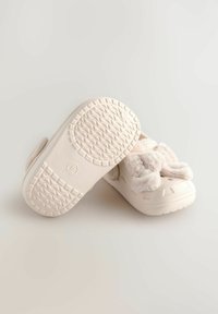 Des chaussures de couleur crème avec une semelle texturée, des accents en fausse fourrure douce et une sangle Velcro sécurisée pour un enfilage facile. Forme arrondie et design respirant.
