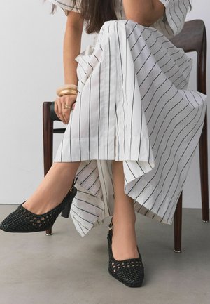 Kobieta siedząca na krześle, ubrana w białą sukienkę w pionowe czarne paski oraz czarne plecione buty na obcasie.