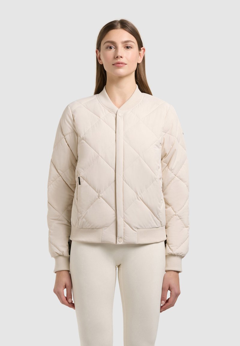 Jeune femme portant une veste bomber matelassée de couleur crème avec des poches avant et un pantalon ajusté assorti, debout devant un fond clair.