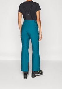 Teal ski pants with adjustable black suspenders, side pockets, and black fabric accents at the hems. Worn with ski boots.