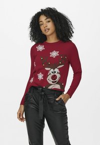 Pull de Noël rouge avec un design de renne souriant, des motifs de flocons de neige et de petites clochettes dorées. Associé à un pantalon en cuir noir.