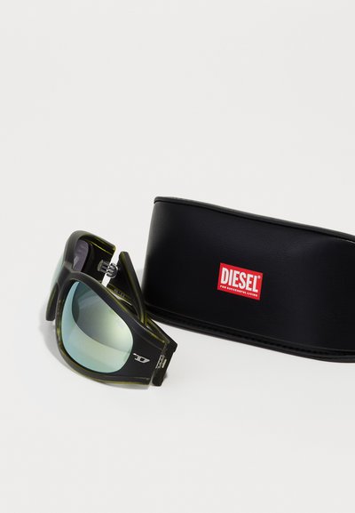 Lunettes de soleil Diesel avec de larges montures noires et vertes, verres dégradés bleus, et un étui noir de marque avec un logo rouge.