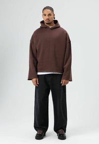Hoodie marron surdimensionné en tissu doux, doté d'un large encolure et de manches longues. Associé à un pantalon noir à jambes larges et des chaussures noires.