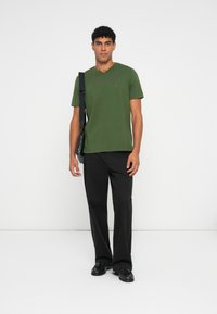 Πράσινη βαμβακερή μπλούζα V με κοντά μανίκια, φορεμένη με μαύρα παντελόνια και παπούτσια. Διαθέτει μικρή λογότυπη λεπτομέρεια στο στήθος.