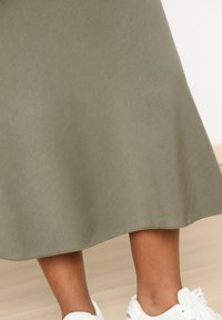 Long, olive green A-line skirt made of smooth fabric with a subtle sheen, featuring a curved hem and paired with white athletic shoes.