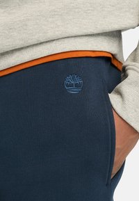 Navy blue cotton sweatpants with a sleek design, featuring an orange waistband and subtle blue embroidered tree logo on the side.