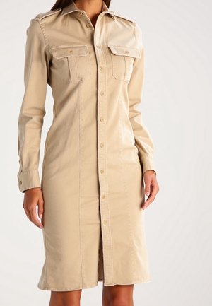 Beige skjortekjole med knapper, lange ærmer, to brystlommer, en søm foran og en midt-længde kant, båret af en stående person.