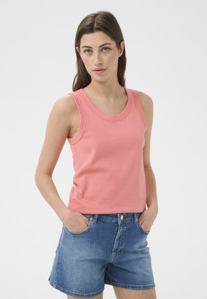 Femme portant un haut sans manches rose et un short en jean bleu, les mains dans les poches, debout devant un fond uni.