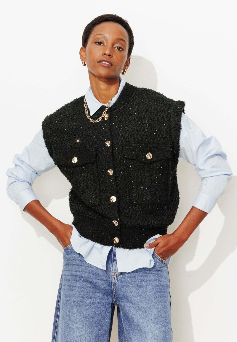 Veste noire texturée avec boutons dorés et poches, portée par-dessus une chemise bleu clair. Modèle portant un jean bleu.