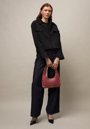 Femme portant une veste courte noire, un jean large foncé, des talons noirs, tenant un sac à main bordeaux clouté sur un fond uni.