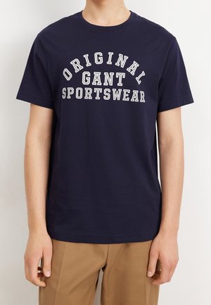 Persoon draagt een donkerblauw t-shirt met witte tekst "ORIGINAL GANT SPORTSWEAR" en lichtbruine broek tegen een egale achtergrond.