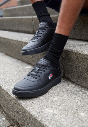 Osoba nosząca czarne trampki Tommy Jeans i czarne skarpetki, siedząca na zewnętrznych betonowych schodach.