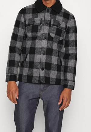 Mann trägt eine graue und schwarze kariertes Wolljacke mit Knopfleiste und Klappentaschen auf der Brust, kombiniert mit dunkelblauen Jeans.