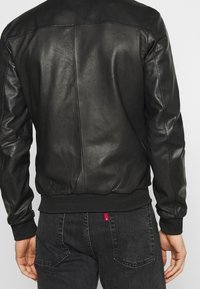 Man i svart läderbomberjacka och mörka jeans som står med ryggen mot kameran mot en enhetlig bakgrund.