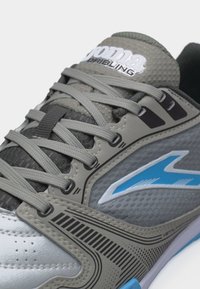Gros plan sur une chaussure de sport grise et bleue avec des lacets, une tige texturée, et un logo de marque sur le côté et la languette.
