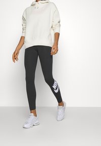 Felpa beige chiaro con cappuccio e lacci, abbinata a leggings neri con il logo Nike bianco. Sneakers grigie completano l'outfit.