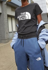Svart bomullströja med vit "THE NORTH FACE"-logotyp. Blå fleecebyxor med elastisk midja och dragsko. Båda har logotypaccent.