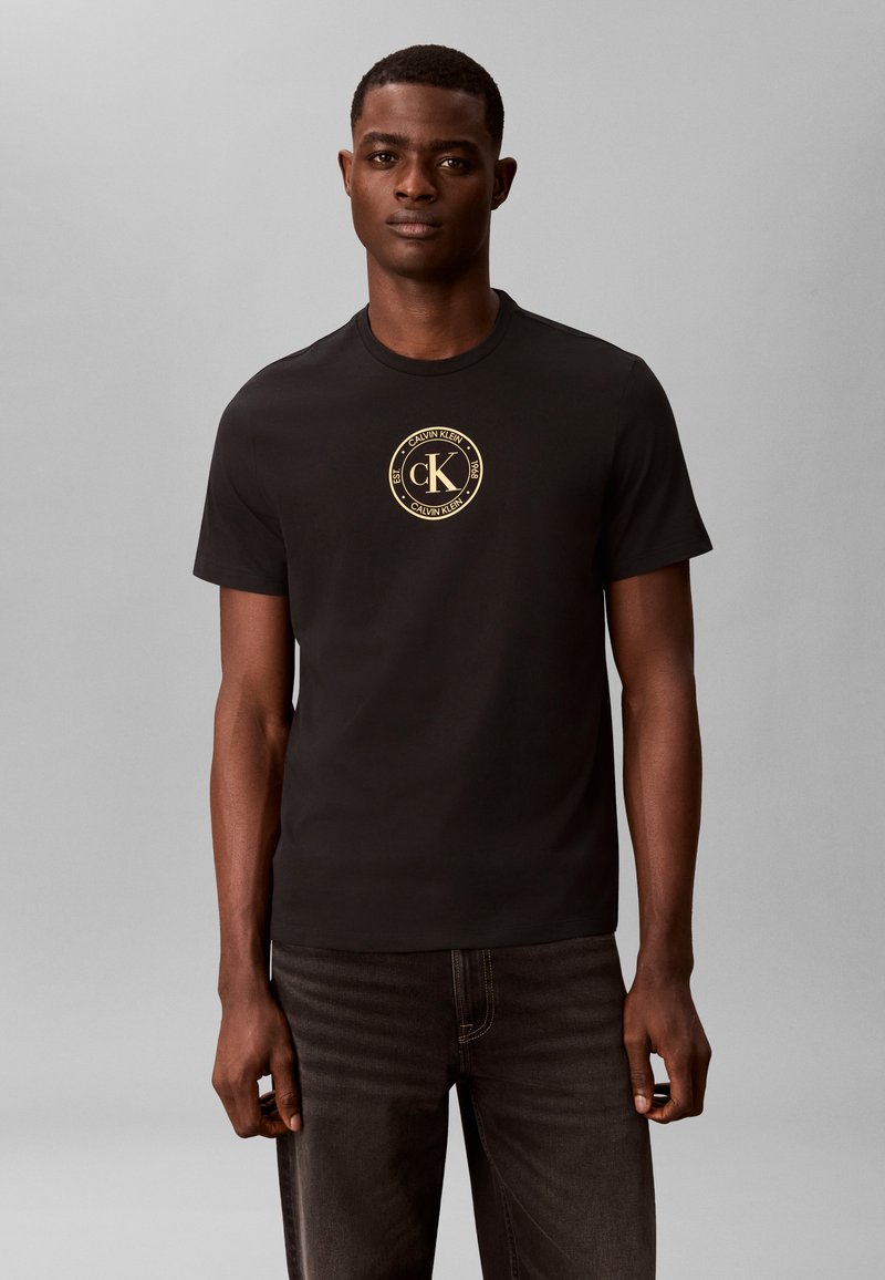 Νεαρός άνδρας φορώντας μαύρο t-shirt Calvin Klein με χρυστό κυκλικό λογότυπο στο στήθος, στέκεται μπροστά σε μια απλή γκρι φόντο.