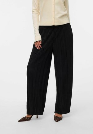 Pantalons larges plissés noirs avec une surface texturée, associés à un cardigan ajusté de couleur claire. Présente une silhouette décontractée.