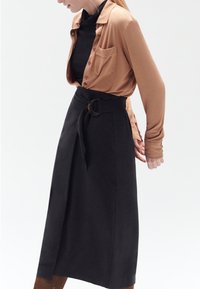 Camicia marrone con bottoni e maniche lunghe, abbinata a una gonna avvolgente nera a vita alta con una grande fibbia circolare.