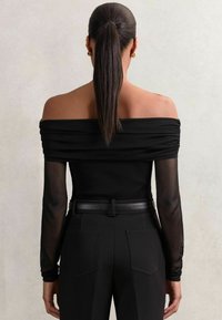 Top negru cu umeri descoperiți, din material plisat, cu mâneci lungi din tul, asortat cu pantaloni negri cu talie înaltă și o curea rectangulară.