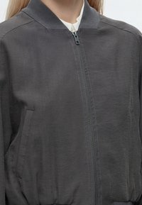 Personne portant une veste zippée gris foncé avec un col côtelé et des poches latérales en biais, visible des épaules à la taille.