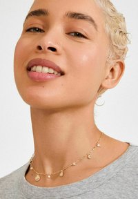 Gouden chokerketting met zon-, maan- en parelhangers, een fijne ketting en gemengde texturen, gedragen rond de halslijn.