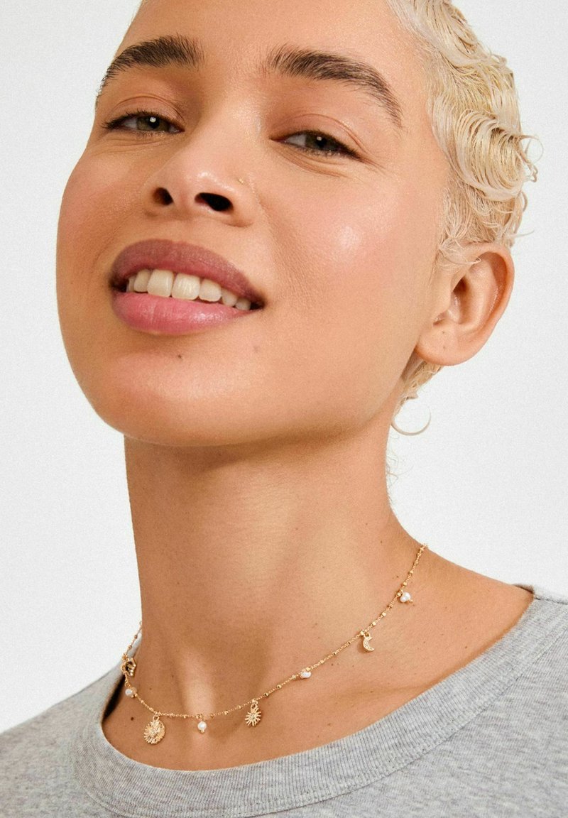 Gouden chokerketting met zon-, maan- en parelhangers, een fijne ketting en gemengde texturen, gedragen rond de halslijn.