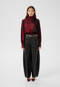 Top lungo a maniche lunghe in raso rosso con colletto alto, abbinato a pantaloni larghi in pelle nera e una cintura nera con borchie argentate, completato da tacchi bordeaux.