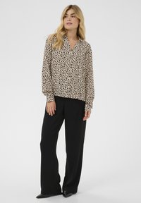 Pikkade blondide juustega naine, kes kannab beežikat leopardimustriga pluusi ja musta laiade säärtega pükse, seisab ühevärvilise heleda tausta ees.