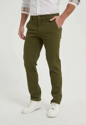 Homme portant un pantalon slim vert olive, des baskets blanches et une chemise blanche aux manches retroussées avec des poignets à carreaux, debout avec une main dans la poche.
