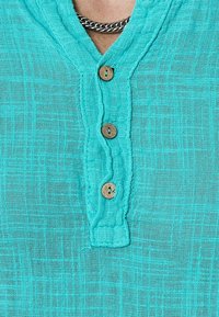 Chemise boutonnée turquoise en tissu léger et texturé. Elle comporte trois boutons en bois au niveau de l'encolure et un col au design décontracté.